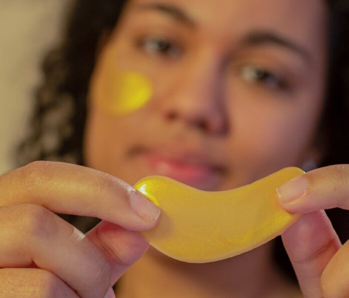 Soft focus of African American young female demonstrating eye patch in hands and looking at camera during skincare routine on beige background